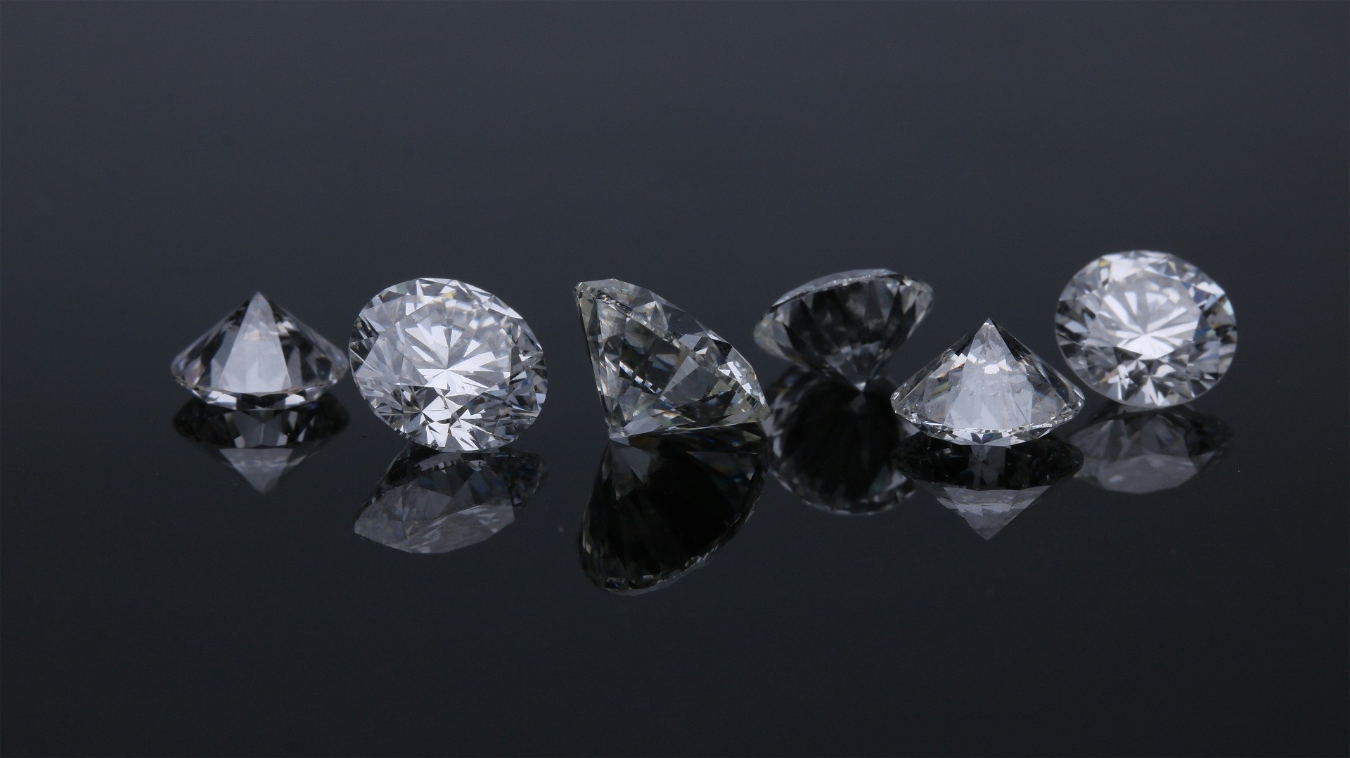 A row of natural round brilliant diamonds arranged on a dark reflective surface, each catching light differently