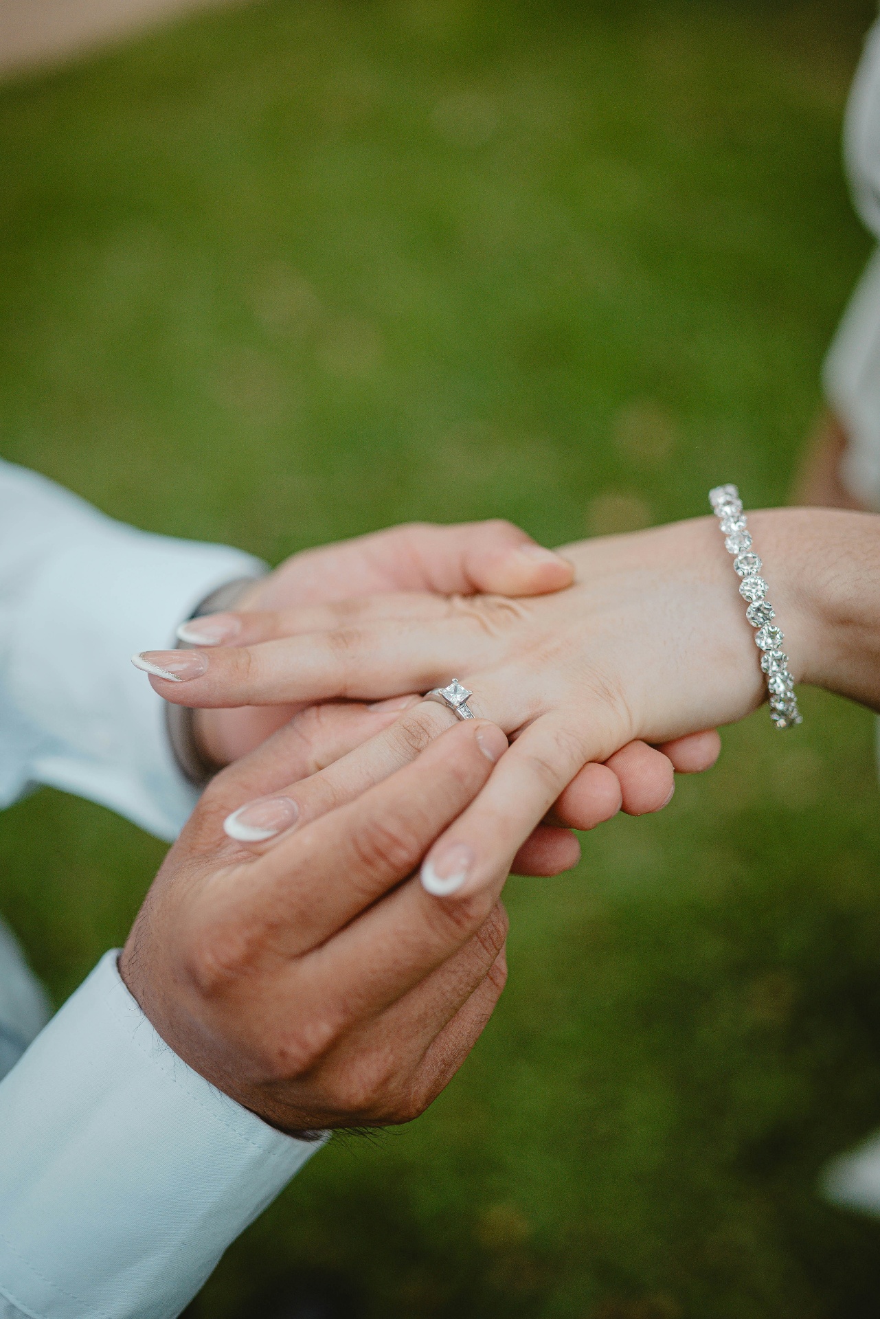 A groom's hands sliding a diamond engagement ring onto his bride's finger, her diamond tennis bracelet catching the light
